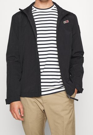 Mand iført sort lynlåsjakke over hvid skjorte med sorte vandrette striber og beige bukser, hænder afslappet ned langs siden.