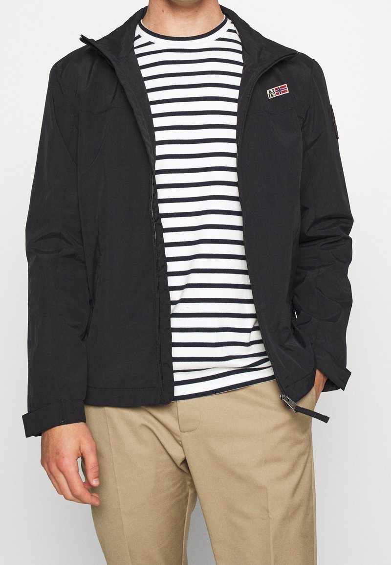 Mężczyzna ubrany w czarną kurtkę na suwak na białą koszulę w czarne poziome paski oraz beżowe spodnie, ręce luźno opuszczone wzdłuż ciała.