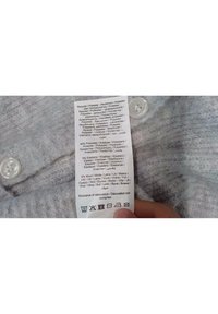 Šedá textilní etiketa s pokyny v několika jazycích, obsahující symboly pro praní, složení materiálu a vedle ní dvě bílé knoflíky.