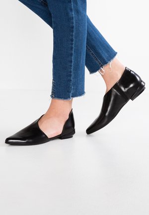 Nohy v krátkých modrých džínách s černými koženými balerínkami s špičatou špičkou a otevřenými boky stojící na bílém podlaze.