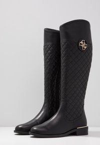 Mustat polvimittaiset saappaat, jotka on valmistettu nahasta ja joissa on tikattu muotoilu. Sisältää kultaisen logon detaljin ja matalan korot. Sujuva pinta ja pyöreä kärki.