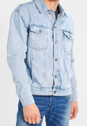Homme portant une veste en jean bleu clair avec fermeture à boutons sur le devant et poches à rabat sur la poitrine, associée à un jean bleu.