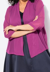 Kvinna i en genomskinlig lila shrug över en svart klänning, med armarna korsade, framför en vit bakgrund.