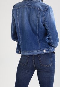 Blå denimjacka med strukturerad design, som har en spetsig krage, knäppning med knappar och en sliten tygtextur.