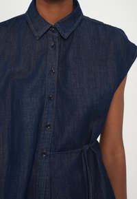 Denimkjole med krageutringning, korte ermer, frontlukking med knapper og belte i livet. Mørk blå med synlige søm detaljer.