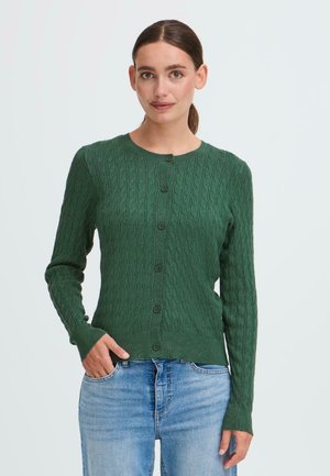 Cardigan en tricot torsadé vert en tissu doux, doté d'un col rond, d'une fermeture à boutons et de manches longues. Porté avec un jean en denim.