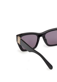 Gafas de sol negras en forma de ojo de gato con lentes morados tintados. Cuentan con un acabado brillante y un acento dorado en la patilla con una marca grabada.