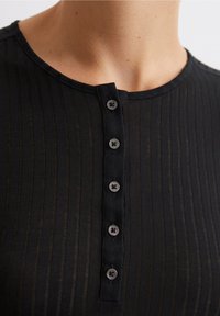 Top nero a coste con scollatura rotonda e placket con sei bottoni in grigio scuro. Il tessuto appare leggero e leggermente trasparente.