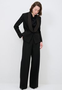 Un costume noir sur mesure avec un revers en satin et un pantalon large. Le tissu a une texture lisse, complété par un blouse noire drapée en dessous.
