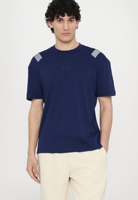 T-shirt en coton bleu marine à manches courtes, avec des accents rayés blancs sur les épaules et un logo EA7 subtil à l'avant.