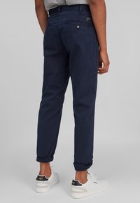 Osoba ubrana w granatowe, podwinięte spodnie casualowe i białe trampki z czarnymi i żółtymi detalami, stojąca na tle jednolitego szarego koloru.