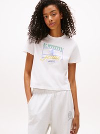 Jeune femme aux cheveux bouclés portant un t-shirt cropped blanc de Tommy Jeans et un pantalon de survêtement gris clair, les mains dans les poches.