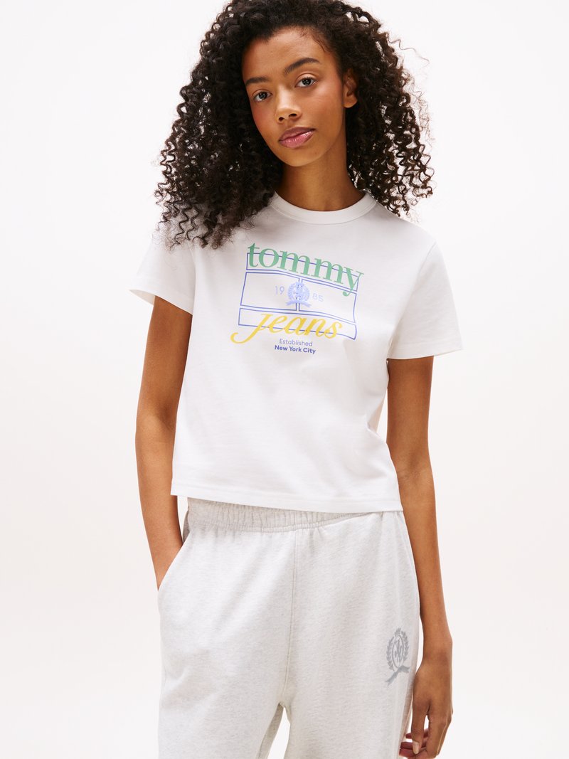 Jeune femme aux cheveux bouclés portant un t-shirt cropped blanc de Tommy Jeans et un pantalon de survêtement gris clair, les mains dans les poches.