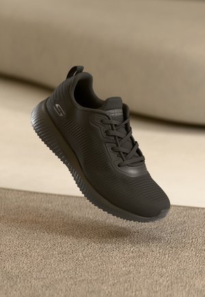 Schwarzer Skechers Sneaker mit strukturiertem Stoff und dicker Sohle, in der Luft über einem beigen Teppichboden dargestellt.