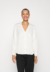 Blusa bianca a maniche lunghe con scollatura a V, chiusura con bottoni frontale e maniche con polsini, realizzata in un tessuto morbido e leggero e con una vestibilità rilassata.