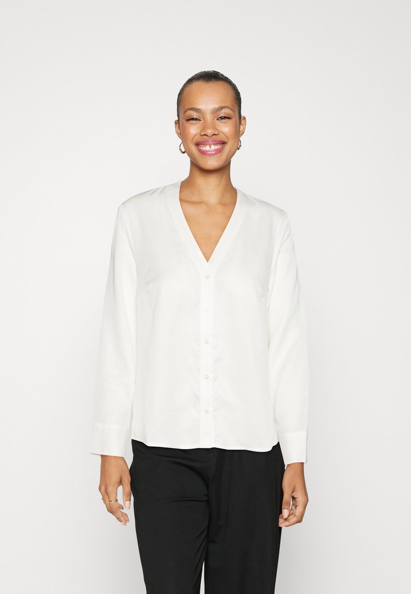 Blusa bianca a maniche lunghe con scollatura a V, chiusura con bottoni frontale e maniche con polsini, realizzata in un tessuto morbido e leggero e con una vestibilità rilassata.