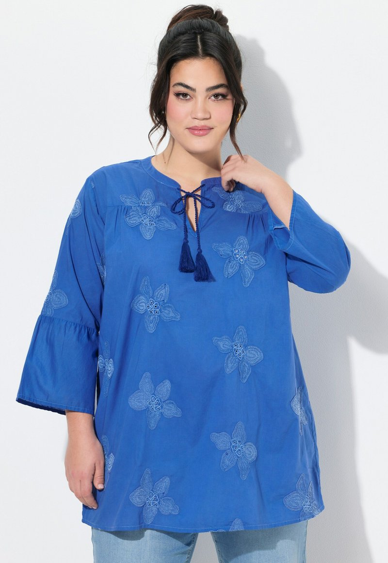 Modrá tunika s květinovou výšivkou, kulatým výstřihem a detailem na zavázání. Má 3/4 zvonové rukávy a měkkou, texturovanou látku. Volný střih.
