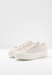 Un paio di sneakers platform bianco sporco con occhielli dorati, allacciatura frontale e bordi ondulati su una superficie bianca.