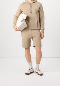 Homme portant un sweat à capuche beige et un short, tenant un sac fourre-tout beige, avec des chaussettes blanches et des baskets blanches à semelles marron.