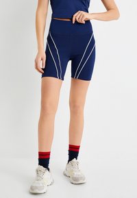 Pantalones cortos deportivos azul marino con ribete diagonal blanco, cintura alta y tejido elástico. Combinados con zapatillas blancas y calcetas a rayas en azul y rojo.