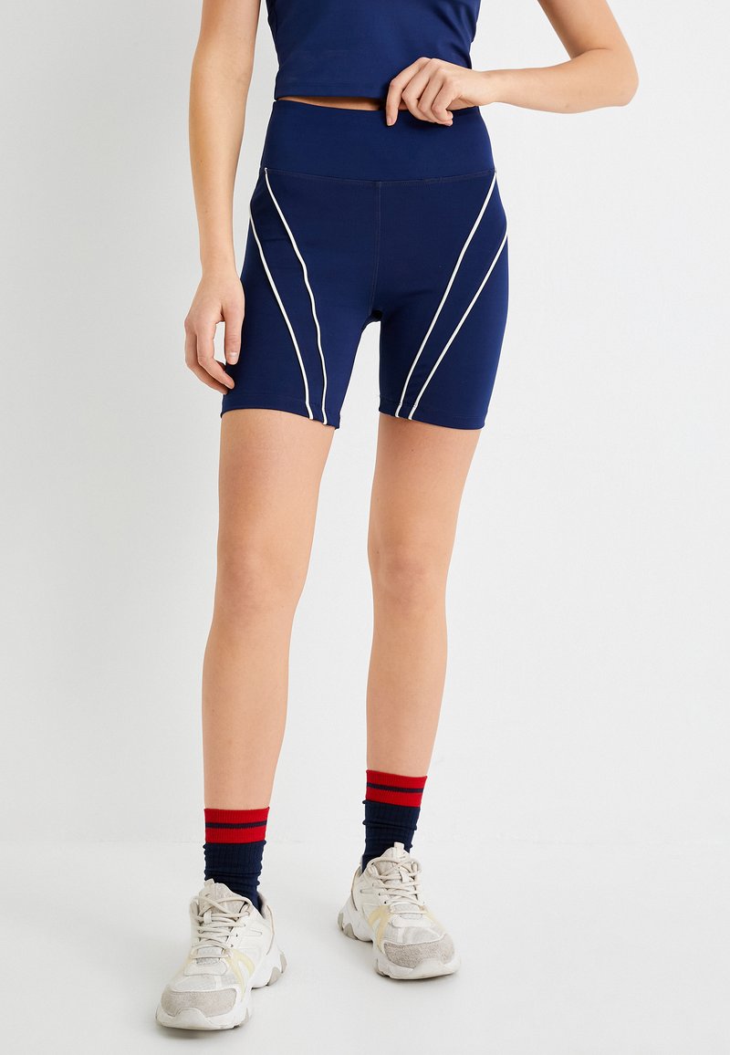 Pantalones cortos deportivos azul marino con ribete diagonal blanco, cintura alta y tejido elástico. Combinados con zapatillas blancas y calcetas a rayas en azul y rojo.