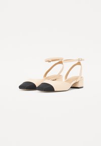 Chaussures à talons bloc en cuir beige avec embouts noirs et brides de cheville. Texture lisse avec un bout arrondi et un design minimaliste.