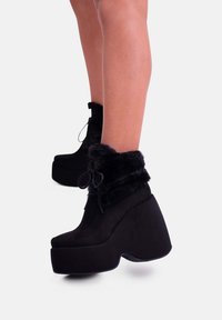 Stivaletti neri con zeppa in suede, dotati di un risvolto in pelliccia sintetica e lacci in raso, con tacco spesso e punta arrotondata.