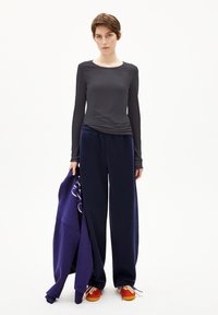 Langärmliges, dunkelgraues Oberteil kombiniert mit navyblauen Jogginghosen; hält eine lila Jacke; mehrfarbige Sneakers mit roten, orangefarbenen und blauen Akzenten.