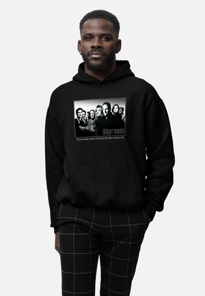 Hombre que lleva una sudadera negra con capucha con foto en blanco y negro del reparto de Sopranos y cita, combinada con pantalones negros con líneas de cuadrícula blancas.