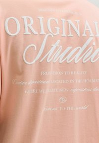 Ροζ βαμβακερό T-shirt με λευκό κείμενο. Το κείμενο περιλαμβάνει τις φράσεις "DESIGNED IN COPENHAGEN" και "ORIGINAL STUDIO" σε διάφορες γραμματοσειρές.