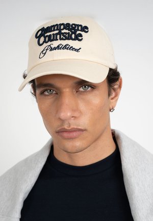 Giovane uomo con cappellino beige chiaro ricamato con la scritta "Champagne Courtside Prohibited", indossa una camicia nera e una giacca grigia sulle spalle, sfondo neutro.