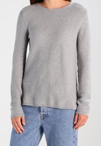Grauer gestrickter Langarmpullover mit rundem Halsausschnitt und gerippter Textur, kombiniert mit hellblauen hochgeschnittenen Jeans. Dezentes Markenlogo am Saum.