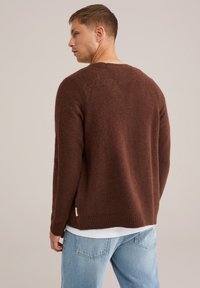 Suéter marrón texturizado con cuello redondo, puños acanalados y un dobladillo trasero ligeramente más largo. Llevado sobre una camiseta blanca con jeans de color azul claro.