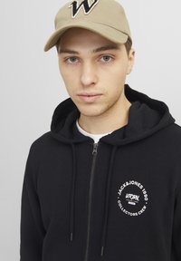 Sudadera negra con cremallera, bolsillo frontal, cordones y un logotipo circular blanco. Combinada con una gorra Beige que presenta una "W" negra.