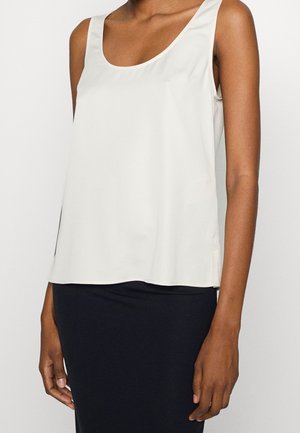 Biała bezrękawnikowa bluzka z zaokrąglonym dekoltem noszona przez osobę o ciemnej karnacji, zestawiona z czarną dopasowaną spódnicą.