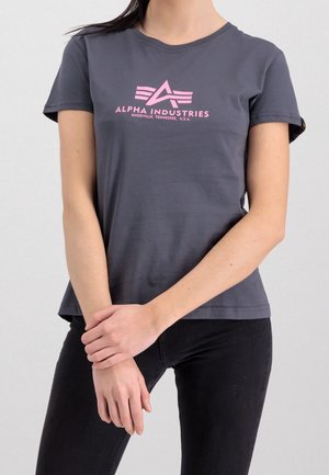 Frau trägt ein dunkelgraues Alpha Industries T-Shirt mit pinkem Logo und Text, kombiniert mit schwarzen Hosen, steht mit vor der Brust verschränkten Armen.