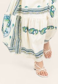 Weißes Kleid mit grünen und blauen Blumenmustern, mit einem ausgestellten Saum. Kombiniert mit beigen Sandalen mit Riemen.