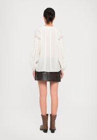 Femme aux cheveux foncés en chignon, portant un haut en dentelle blanche, une mini-jupe en cuir noir et des bottes à la cheville marron, se tenant de dos sur un fond blanc.