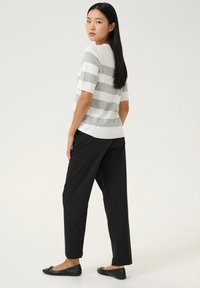 Gestricktes gestreiftes Oberteil in Hellgrau und Weiß, kombiniert mit schwarzen locker sitzenden Hosen und flachen schwarzen Schuhen. Modell steht mit dem Rücken zur Kamera.