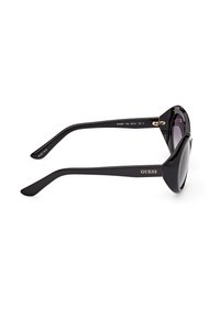 Lunettes de soleil noires surdimensionnées en forme d'œil de chat avec des verres teintés et le logo "GUESS" sur l'arête du côté, vues de profil sur un fond blanc.