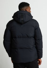 Melnā pūkaina jaka ar kapuci, ar pārklājumu, regulējamiem siksnām un logotipu augšējā muguras daļā. Gluda tekstūra.