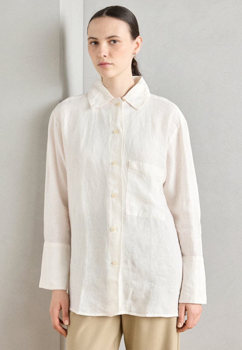 Linen button-up shirt in light cream with a collar, long sleeves, a chest pocket, and a relaxed fit. Textured surface with natural fibers.