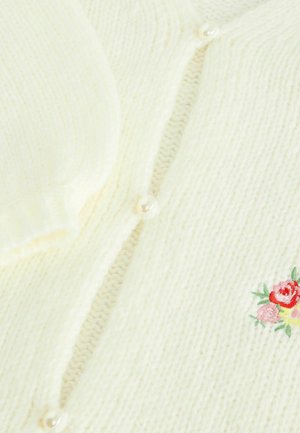 Cream knit fabric with pearl buttons and a small embroidered floral design in red, pink, and green on the lower right side.