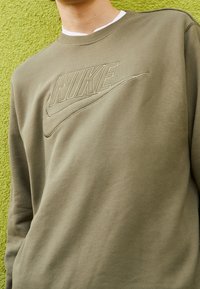Sweatshirt Nike vert olive avec logo en relief, col ras du cou, texture douce et manches longues, sur fond vert vif.