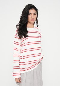 Λευκό πουκάμισο με μακριά μανίκια και οριζόντιες κόκκινες ρίγες. Το σχέδιο έχει χαλαρή εφαρμογή και στρογγυλή λαιμόκοψη, συνδυασμένο με πλισέ γκρι φούστα.