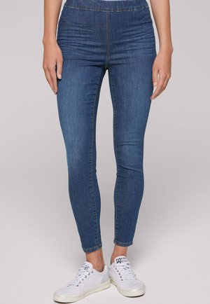 Woman wearing blue skinny jeans and white sneakers standing against a plain light background.