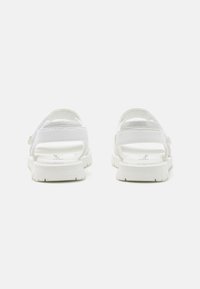 Białe sandały z teksturowanym paskiem, otwartą piętą i wyściełaną wkładką, wyposażone w gumową podeszwę z lekkim wzorem antypoślizgowym.