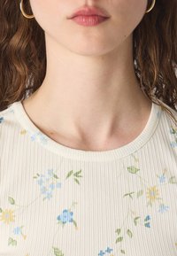 Ραβδωτό κρεμ ύφασμα με λουλουδάτο σχέδιο που περιλαμβάνει μπλε και κίτρινες ανθοσυνθέσεις. Στρογγυλή λαιμόκοψη και απαλή υφή, αναδεικνύοντας τις λεπτές λεπτομέρειες.