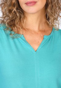 Türkisfarbenes V-Ausschnitt-Oberteil aus weichem Stoff mit glatter Textur. Verfügt über sichtbare Nähte und einen lässigen Schnitt, der den Ausschnitt hervorhebt.