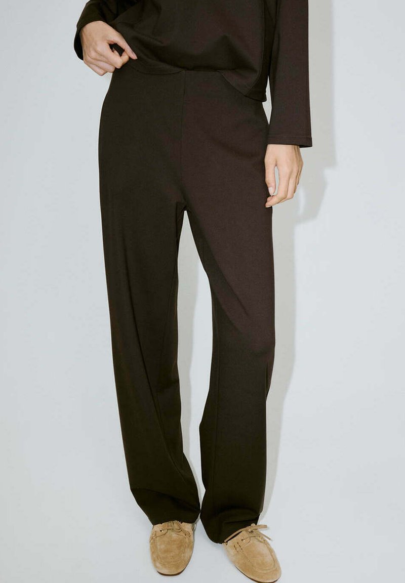 Personne portant un pantalon noir ample et des mocassins en daim beige, debout devant un fond clair uni.